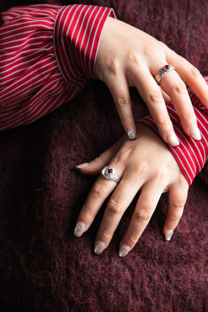 A person with manicured nails wears two silver rings for short fingers, their hands resting on a dark maroon textured fabric while wearing a red and white striped sleeve. earbij